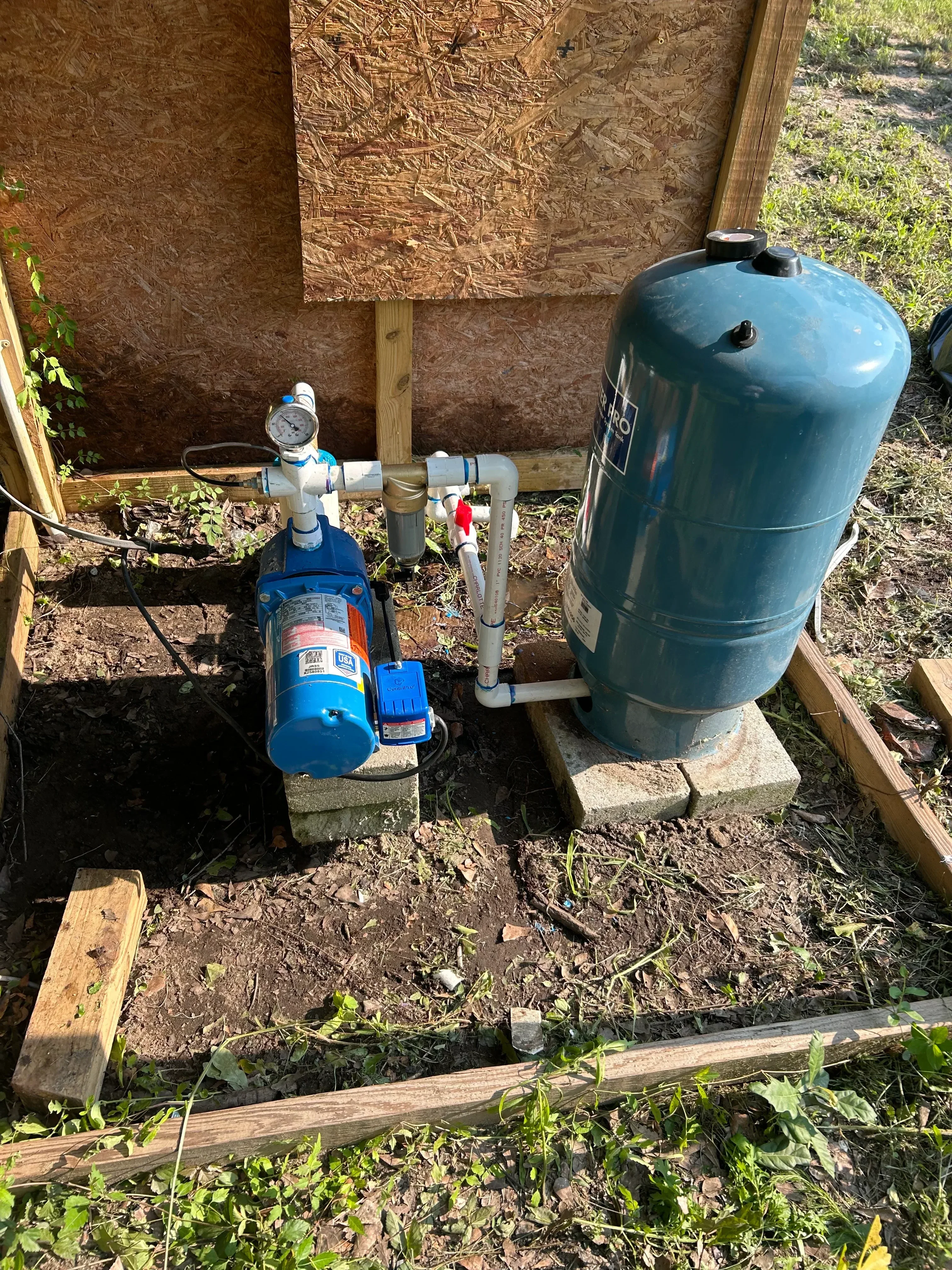Blue pressure tank with pump unit, PVC piping, and pressure gauge mounted on wooden frame