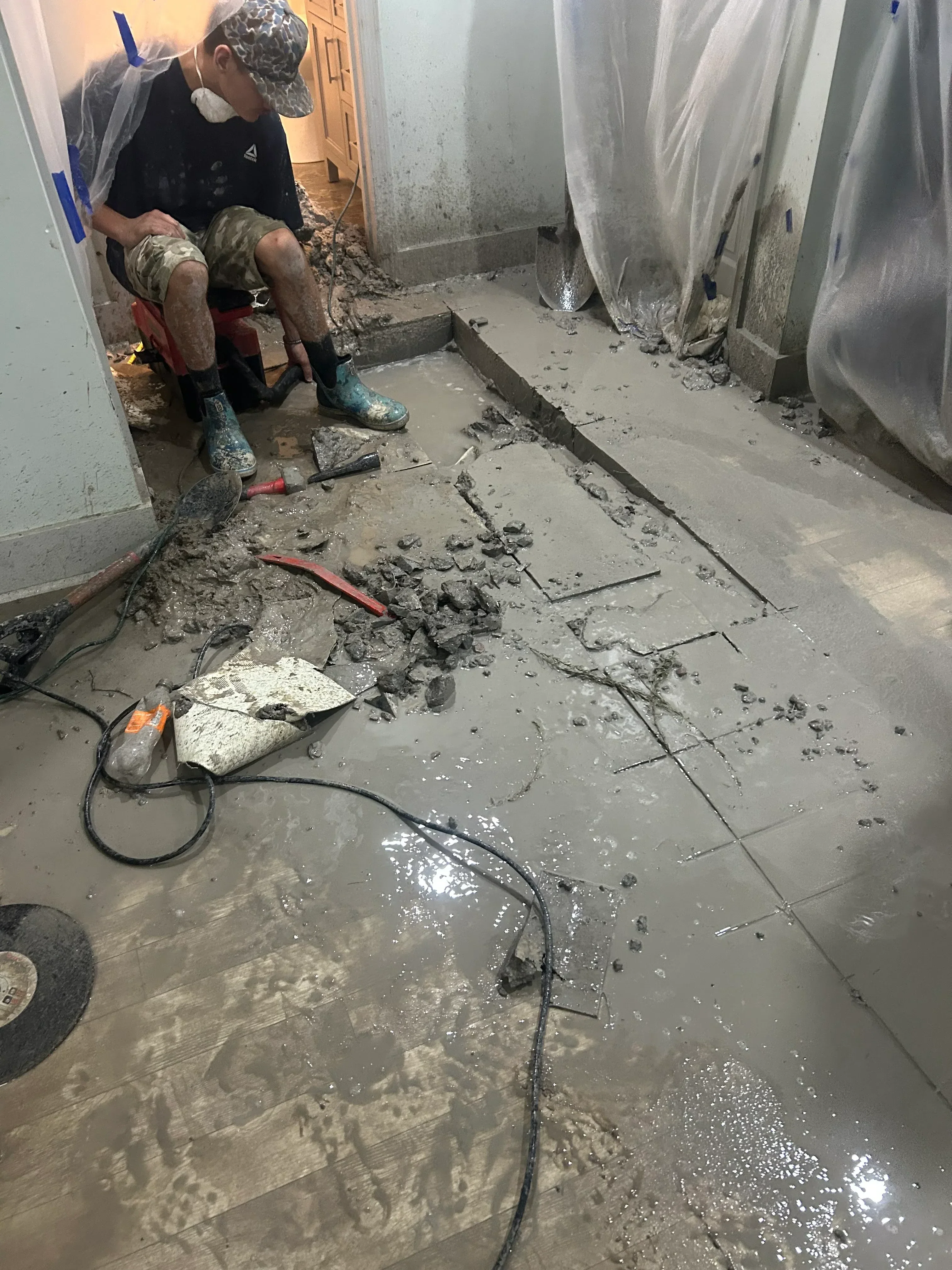 Worker breaking concrete floor with jackhammer to expose plumbing lines and conduit