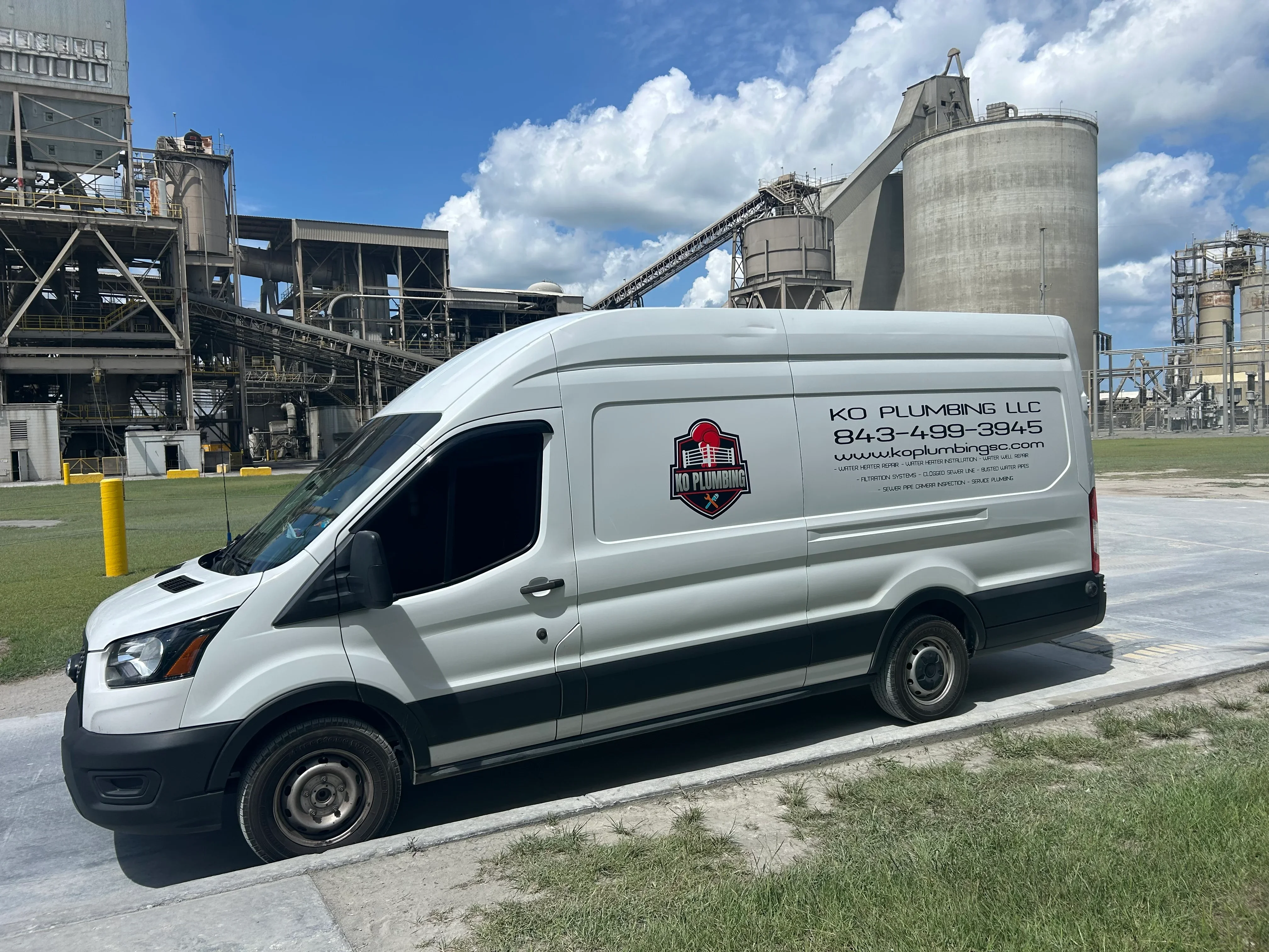 White service van with vinyl wrap branding parked at industrial facility with silos