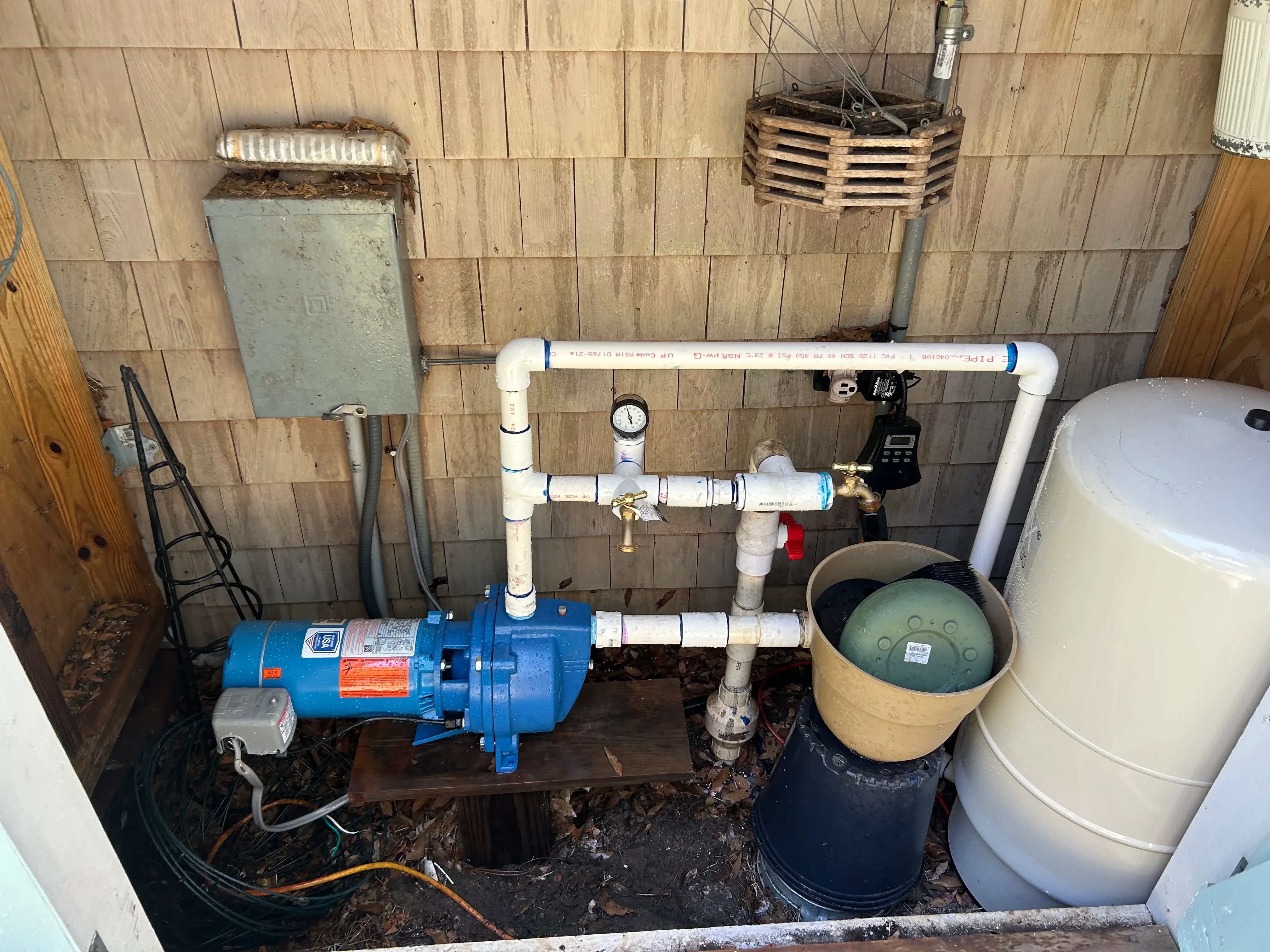 Blue centrifugal well pump with white PVC piping, pressure tank, and gauge on wood foundation
