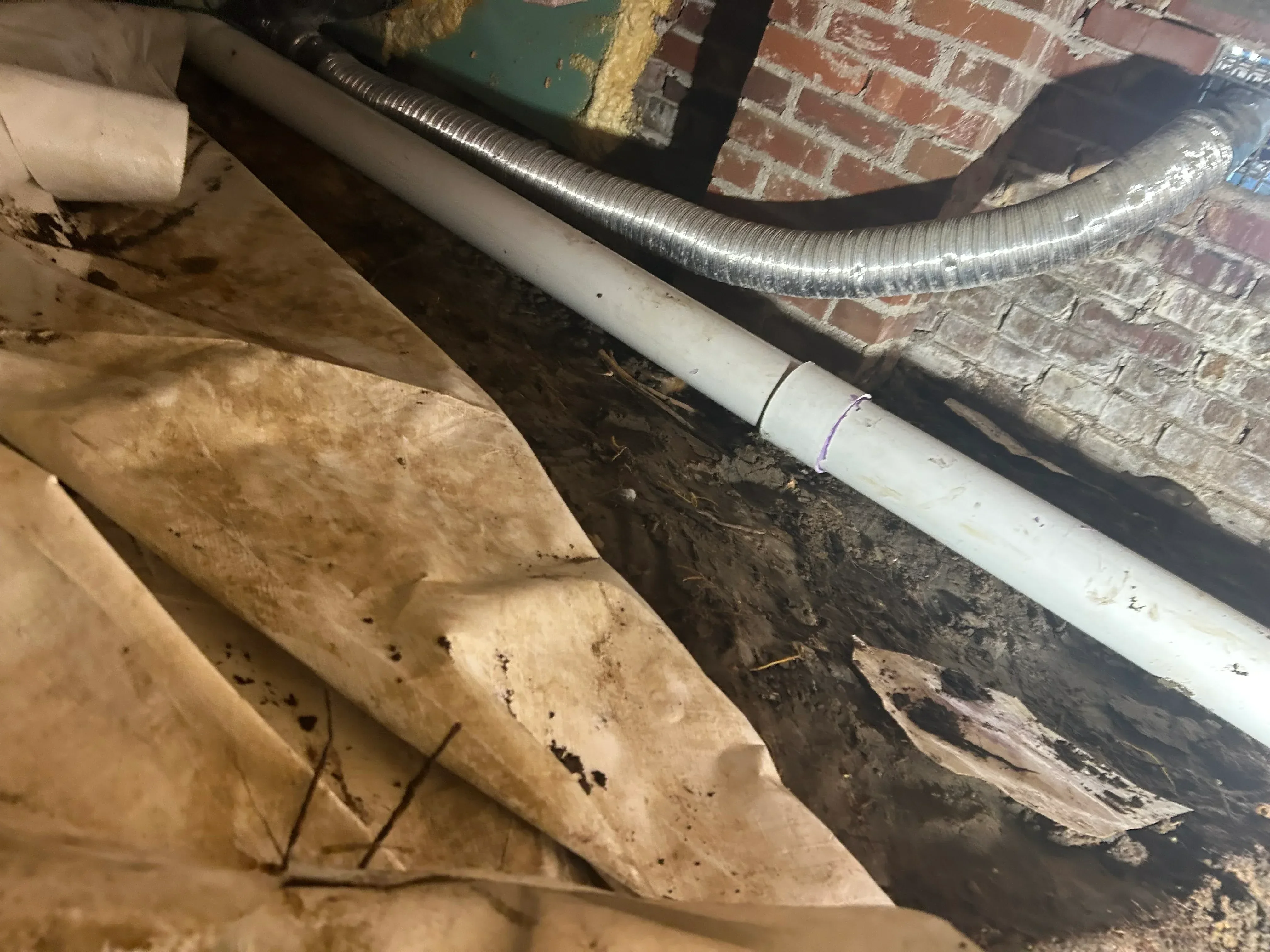 White PVC drain pipe with metal flex duct installed in basement near brick foundation and wooden joists