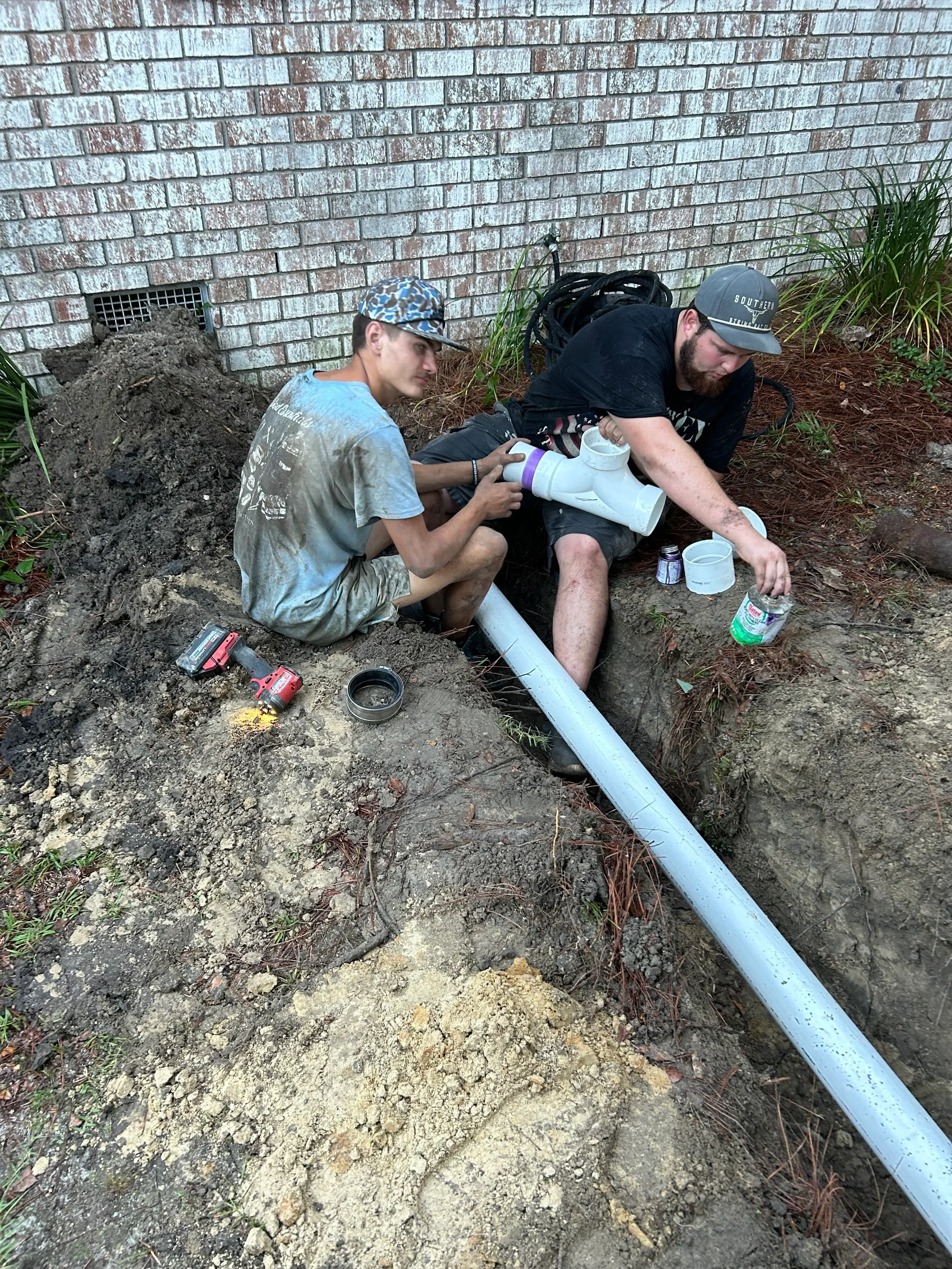 Two plumbers installing blue PVC drain pipe in trench with soil and gravel base