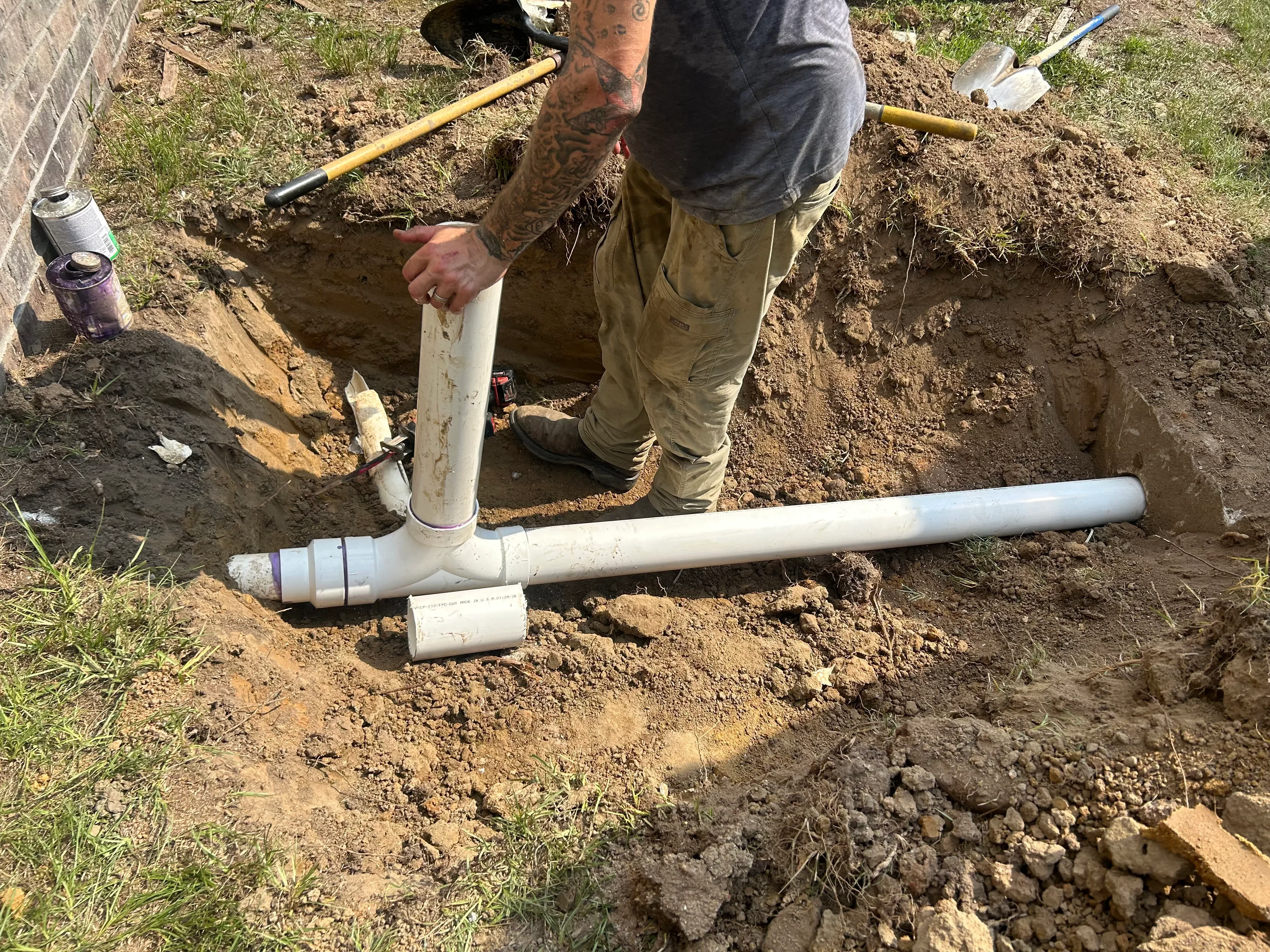 Technician installing white PVC drain pipe with T-junction fitting in excavated trench